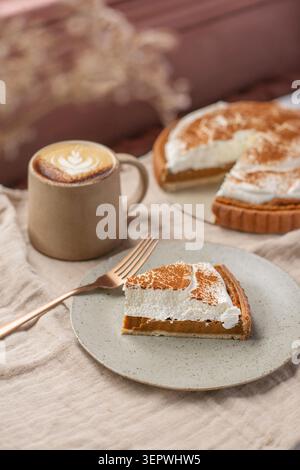 A slice of pumpkin pie topped with whipped cream and cinnamon next to a hot cappuccino with latte art, with the whole pie in the background. Stock Photo