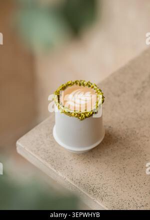 A hot pistachio latte showcasing detailed leaf art in a white ceramic cup with a crushed pistachio rim, set on a textured stone surface. Stock Photo
