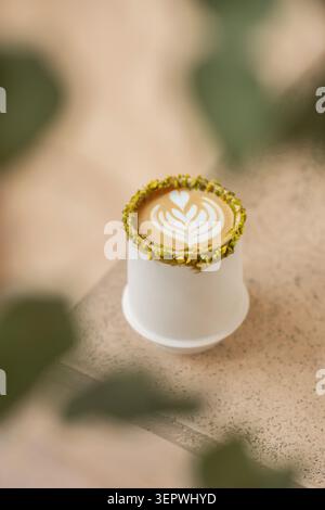 A hot pistachio latte showcasing detailed leaf art in a white ceramic cup with a crushed pistachio rim, set on a textured stone surface. Stock Photo