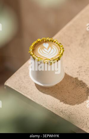 A hot pistachio latte showcasing detailed leaf art in a white ceramic cup with a crushed pistachio rim, set on a textured stone surface. Stock Photo