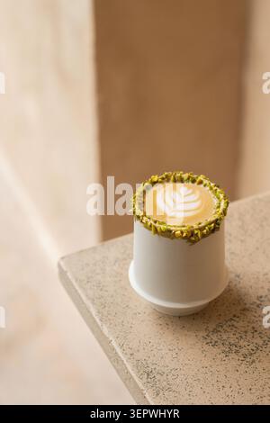 A hot pistachio latte showcasing detailed leaf art in a white ceramic cup with a crushed pistachio rim, set on a textured stone surface. Stock Photo