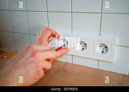 Hand installing electrical outlet socket on kitchen wall, using a screwdriver for home renovation, electrical work, and power supply connection during apartment repair Stock Photo