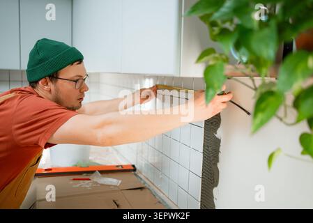 Man installing white ceramic backsplash tiles with adhesive in a modern kitchen, using a measuring tape to ensure precise alignment for a home improvement project Stock Photo