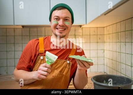 Happy man in a green beanie and overalls smiles at the camera during a home renovation. He holds a spatula with green putty, which is also playfully smeared on his face. Warm, positive mood. Stock Photo