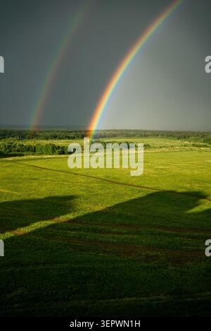 Double rainbow arching over a verdant rural landscape with a stormy sky, casting long shadows on the bright green meadow, creating an inspiring scene of nature's beauty Stock Photo