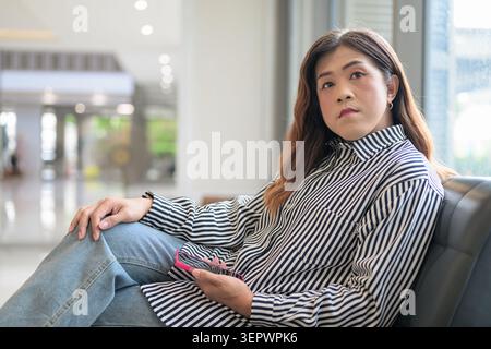 Thai Isan transgender businesswoman in 40s sitting on leather sofa with phone indoors. Horizontal medium shot of a mature non-binary Southeast Asian t Stock Photo