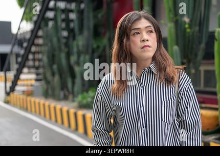 Thai Isan transgender spokeswoman looking aside in pensive thought by street. Horizontal medium shot portrait of a mature non-binary Southeast Asian t Stock Photo