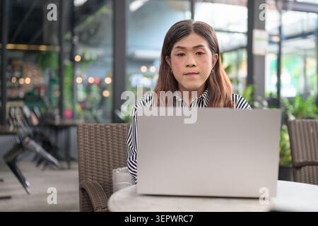 Thai Isan transgender businesswoman in 40s using laptop at cafe terrace. Horizontal medium shot portrait of a mature non-binary Southeast Asian trans Stock Photo