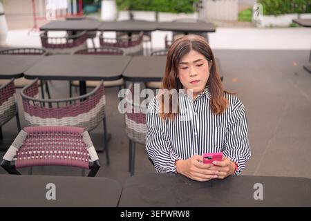 Thai Isan transgender businesswoman in 40s texting on smartphone at cafe veranda. Horizontal medium wide shot of a mature non-binary Southeast Asian t Stock Photo