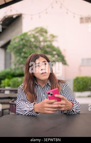 Thai Isan transgender spokeswoman holding smartphone looking up thoughtfully outdoors. Vertical medium shot portrait of a mature non-binary Southeast Stock Photo