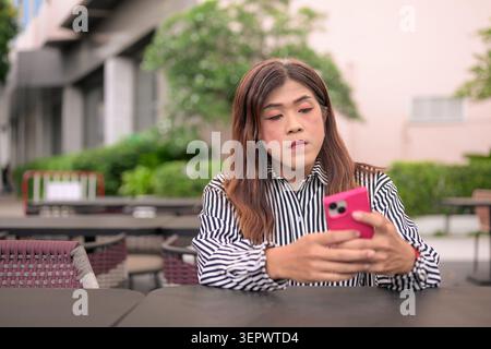 Thai Isan transgender businesswoman in 40s using mobile smartphone at patio table. Horizontal medium shot of a mature non-binary Southeast Asian trans Stock Photo