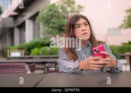 Thai Isan transgender businesswoman in 40s looking away thoughtfully with smartphone. Horizontal medium shot portrait of a mature non-binary Southeast Stock Photo
