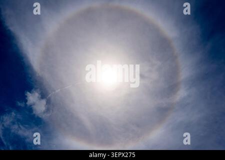Bright sun surrounded by a large circular halo and wispy cirrostratus clouds, creating ethereal light rings and subtle rainbow colors against a deep b Stock Photo