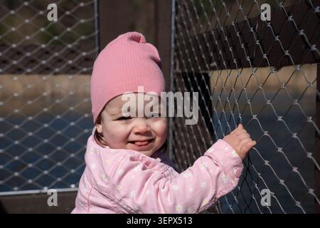 close-up portrait of a smiling toddler wearing a pink knit beanie and daisy-patterned jacket, gripping a diamond-mesh safety fence on a sunlit wooden Stock Photo