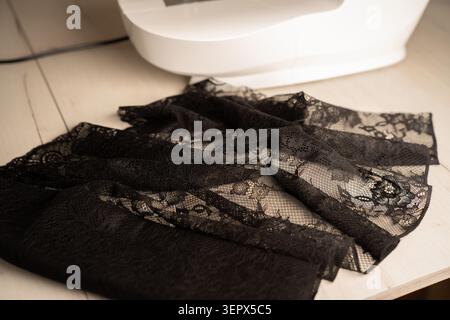 closeup view of black floral lace fabric casually draped over a pale wooden tabletop next to the white sewing machine base, highlighting sheer intrica Stock Photo