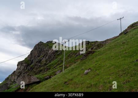 Utility poles and overhead power lines extending across a steep green mountainside beneath dramatic storm clouds. Image representing electrical infras Stock Photo