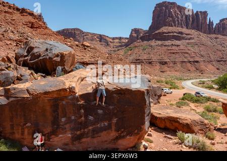 A young male climber tops out bouldering on The Black Box. Big Bend Bouldering Area, Moab, Utah.  A young woman is resting by the boulder. Stock Photo
