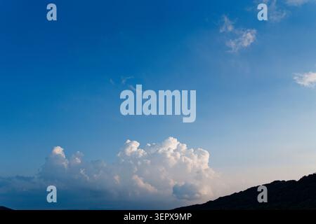 A dark mountain ridge silhouette glows against a vivid deep blue dusk sky with billowing white cumulus clouds on the horizon. Stock Photo