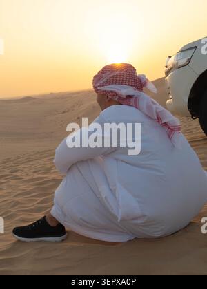 Back view of a man wearing traditional Middle Eastern clothing with a ghutra headscarf, looking out at the horizon during sunset. Stock Photo