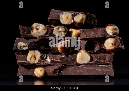 Broken dark chocolate bars stacked with whole hazelnuts on black background. Close up of dark chocolate pieces with whole hazelnuts stacked on top of Stock Photo