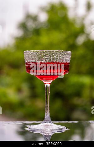 Red cocktail in crystal coupe glass on dark reflective surface outdoors during rain. Close up of vibrant red drink in textured vintage coupe glass aga Stock Photo