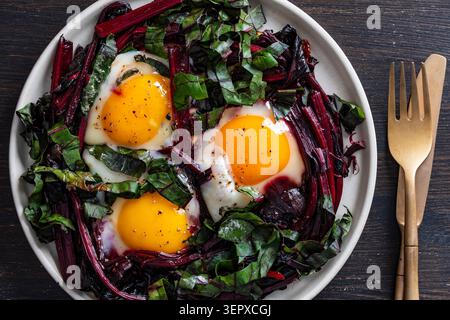 A vibrant, high-angle close-up photograph of three sunny-side up eggs fried with a generous helping of red beet stems and dark green leaves in a white Stock Photo