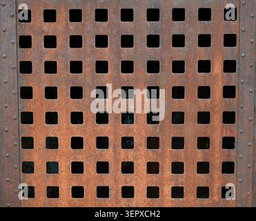 Horizontal close up of a rust coated metal surface with a grid of square cut outs, pitted corrosion, dark openings, and two vertical rows of rivets Stock Photo