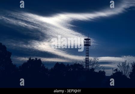 Silhouette of a telecommunications tower and tree line against a dramatic streaky cloudy sky, in Asagiri Kumamoto Japan Stock Photo