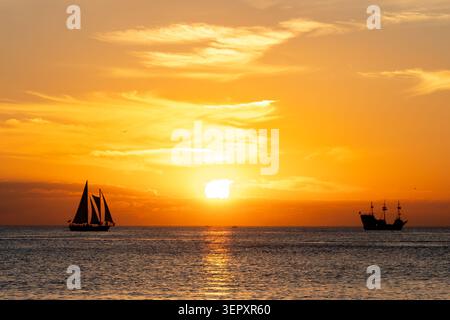 A sunset cruise and small sailboat silhouetted against the setting sun over a calm ocean. Stock Photo