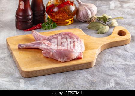 Fresh raw rabbit legs on wooden cutting board with garlic, oil, pepper, and spices on gray stone surface. Closeup of meat preparation for cooking Stock Photo