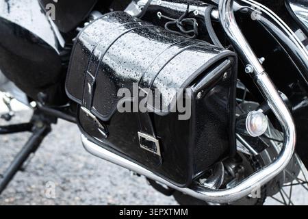 Close up detail of black leather motorcycle saddlebag, water drops visible on surface after rainfall. Chrome elements reflect ambient light, showcasin Stock Photo