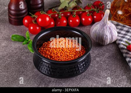 Raw red lentils in black ceramic bowl surrounded by cherry tomatoes, garlic, basil, pepper mill and oil on stone surface Stock Photo