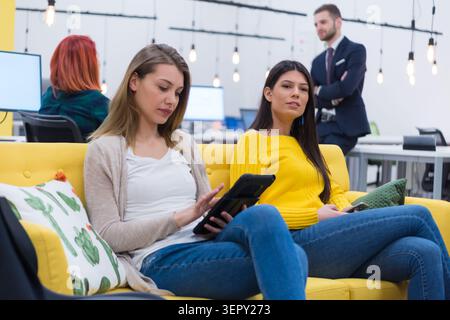 A group of young professionals gathered in a colorful lounge area. Two women sit on a bright yellow sofa using a tablet, while colleagues sit on beanb Stock Photo