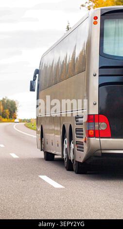 Gold colored coach bus drives away from viewer on winding road. Rear and side view of vehicle show sleek design and multiple wheels. Scenic autumn lan Stock Photo