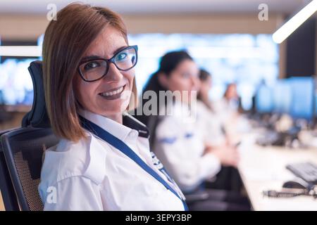 Portrait of a friendly, professional young female security guard wearing glasses and a uniform, smiling at the camera while working with colleagues in Stock Photo