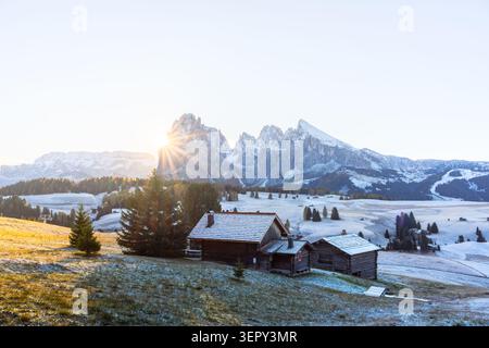 Sunstar over Sassolungo above snow-covered meadows on Alpe di Siusi in the Dolomites. Early winter landscape with traditional mountain huts and frosty grass Stock Photo