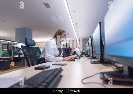 Side profile view of a professional young female security officer in uniform focused on data on multiple computer screens inside a modern surveillance Stock Photo