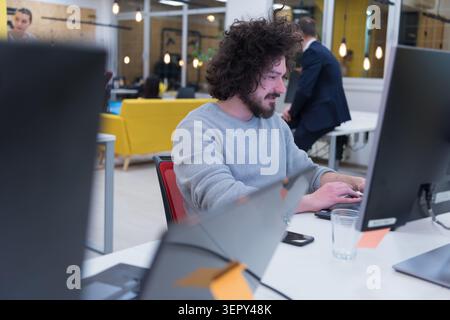 Close-up portrait of a cheerful man with curly hair and a beard, wearing a grey sweater, smiling at the camera while working at a computer in a bustli Stock Photo