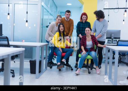 A playful group of young professionals engage in a lighthearted moment, pushing colleagues on rolling office chairs through a modern open-plan office. Stock Photo