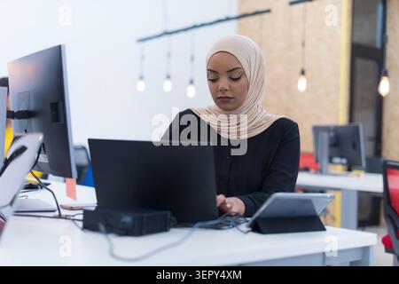 Two young women in hijabs pose for a portrait in a stylish office featuring industrial lighting, with a collaborative team visible behind them. Stock Photo