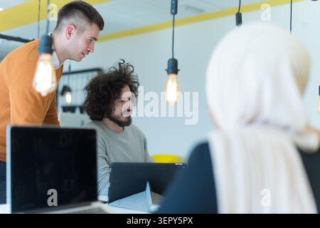 A collaborative business meeting featuring a young man standing and gesturing toward a male colleague with curly hair who is looking at a tablet. In t Stock Photo