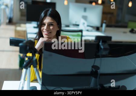 A young woman with long dark hair leans on her hand and smiles warmly toward the camera from behind her desktop computer setup. The blurred background Stock Photo
