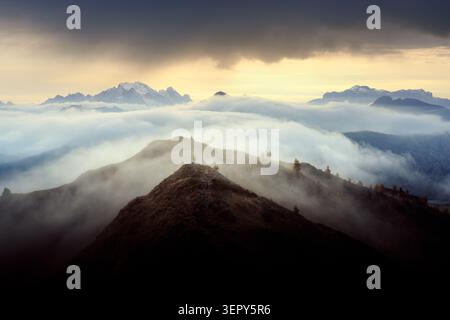 A lone hiker stands above a sea of clouds near Passo Giau at sunrise in the Dolomites. Dramatic mountain landscape with misty valleys, layered ridges and golden light Stock Photo