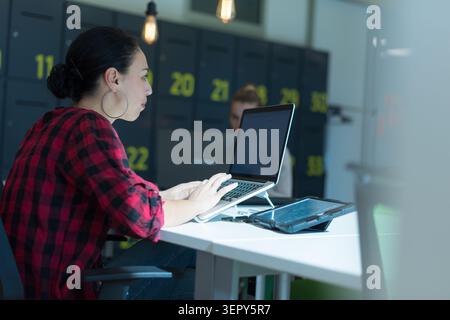 Side profile of a young woman with dark hair tied back in a bun, wearing a red and black plaid shirt, typing intently on her laptop. Her workstation i Stock Photo