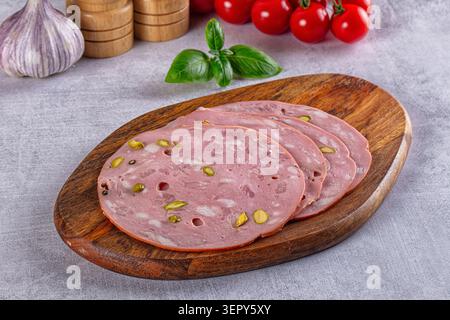 Thinly sliced pink mortadella sausage studded with green pistachios arranged on oval wooden cutting board, accompanied by fresh basil leaves, garlic b Stock Photo