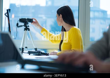 A young female blogger or content creator in a bright yellow sweater adjusts the screen of a digital video camera mounted on a tripod at her desk. She Stock Photo