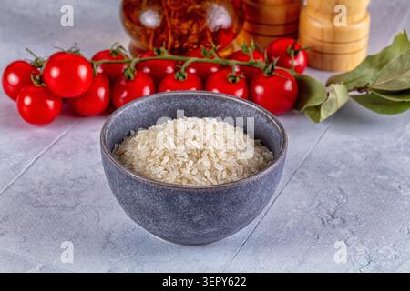 Raw white rice in ceramic bowl with red cherry tomatoes on vine, bay leaves and spices. Healthy cooking ingredients on light stone background Stock Photo