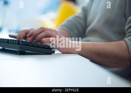 A shallow depth of field shot focusing on a man's hands typing on a black mechanical computer keyboard at an office desk. A colleague in a yellow swea Stock Photo