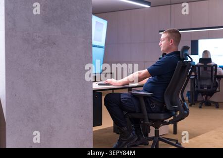 Side profile of a male security officer sitting in an ergonomic office chair, using a keyboard and tracking real-time information across stacked dual- Stock Photo