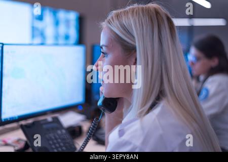 A close-up profile shot of a blonde female security operator talking on a desk phone, with computer screens displaying maps and data visible in the ba Stock Photo